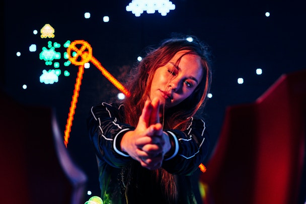 A woman with her fingers in the shape of a gun infront of gaming-style neon signs