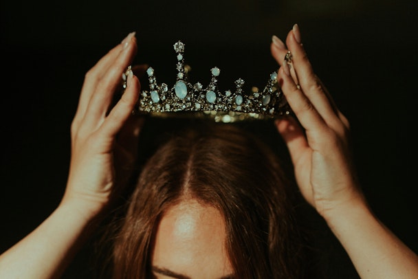 A person with long brown hair holds a crown over their own head