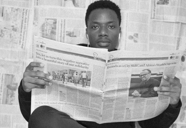 A man reads a newspaper while sitting in front of a wall covered in newspapers