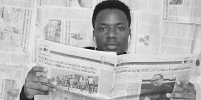 A man reads a newspaper while sitting in front of a wall covered in newspapers